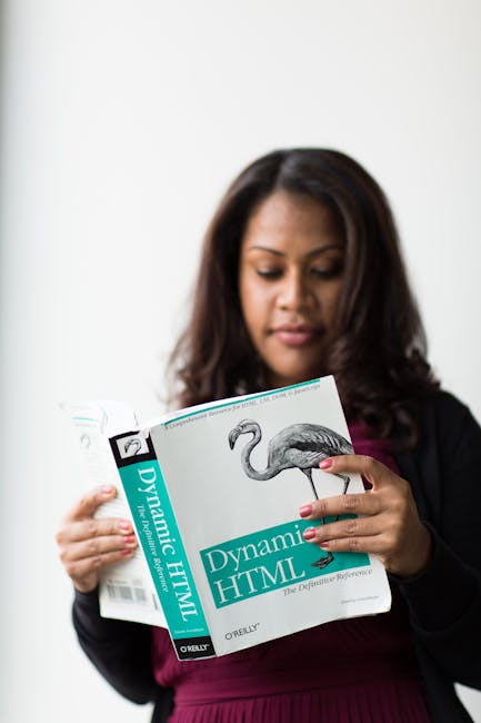 The image shows a woman with shoulder-length dark hair reading a book titled 'Dynamic HTML' published by O'Reilly. She is dressed in a black blazer over a burgundy top, standing against a plain, light