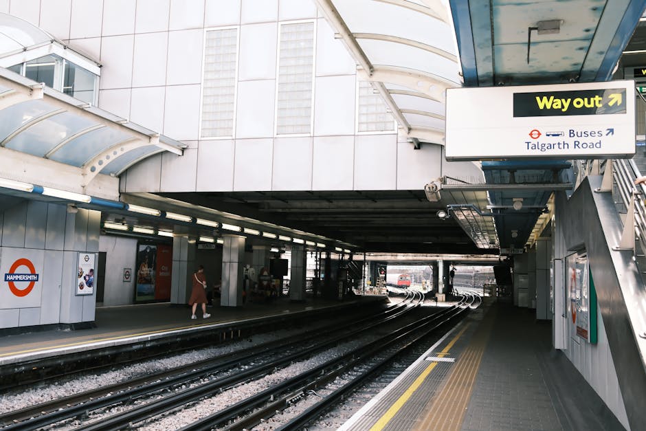 An underground train station platform located beneath a modern building, featuring multiple railway tracks curving into the distance. The platform has a textured, slip-resistant surface with yellow sa