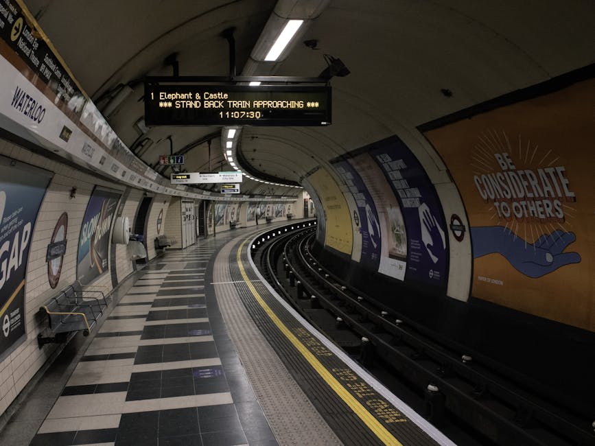 An underground train station platform located beneath a modern building, featuring multiple railway tracks curving into the distance. The platform has a textured, slip-resistant surface with yellow sa