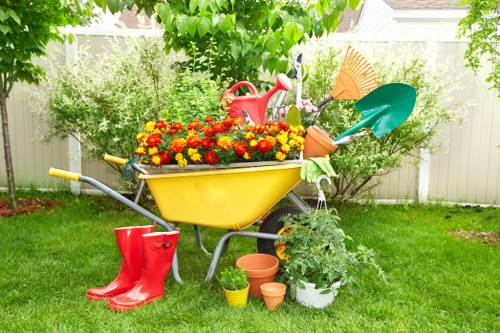A vibrant garden scene featuring a lush, well-maintained lawn with a foreground of green grass and a white fence in the background. Central to the image is a yellow wheelbarrow filled with an abundance of bright red and yellow flowering plants, sitting on a partly shaded lawn. Surrounding the wheelbarrow are various gardening tools and accessories, including a red watering can resting inside the wheelbarrow, a pair of red rubber boots on the ground, and several small, empty terracotta and plastic plant pots placed nearby. To the right, a potted shrub with variegated green and white leaves extends over the ground, and a green garden hose is coiled partially visible behind the wheelbarrow. The backdrop captures an overcast or partly cloudy sky, contributing diffused natural light that enhances the natural earthy tones of the garden elements, reflecting a tidy, actively maintained outdoor space suitable for professional gardening and lawn care services in Uxbridge.