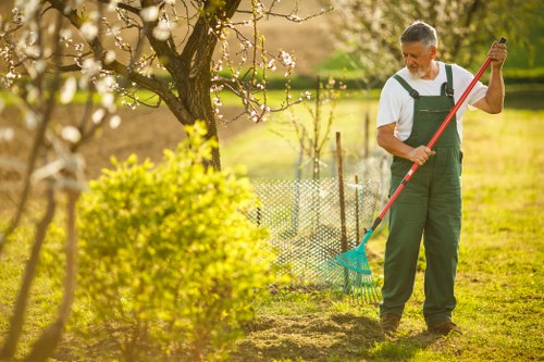 A man in gardening attire, including a white t-shirt and dark overalls, is working in a well-maintained garden in Uxbridge. He is using a rake with a long red handle to tend to the soil area, which is partially shaded by a large tree with budding branches. The garden features a lush green lawn with neatly trimmed grass and a small flowerbed bordered by a wooden edge, displaying mixed plants and young shrubs. In the background, there is a wire mesh fence separating the garden from an open grassy field, indicating a spacious outdoor environment typical of a suburban garden in Uxbridge. The scene is illuminated by warm sunlight, suggesting a pleasant spring or summer day. The absence of other tools or equipment emphasizes a focus on general maintenance and lawn care, aligning with professional gardening services provided by Garden Maintenance Uxbridge.